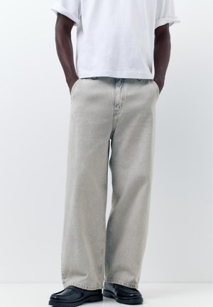 Person trägt hellgraue Jeans mit weitem Bein, ein weißes Hemd mit hochgekrempelten Ärmeln, schwarze Loafer, mit Händen in den Taschen, stehend vor einem schlichten Hintergrund.