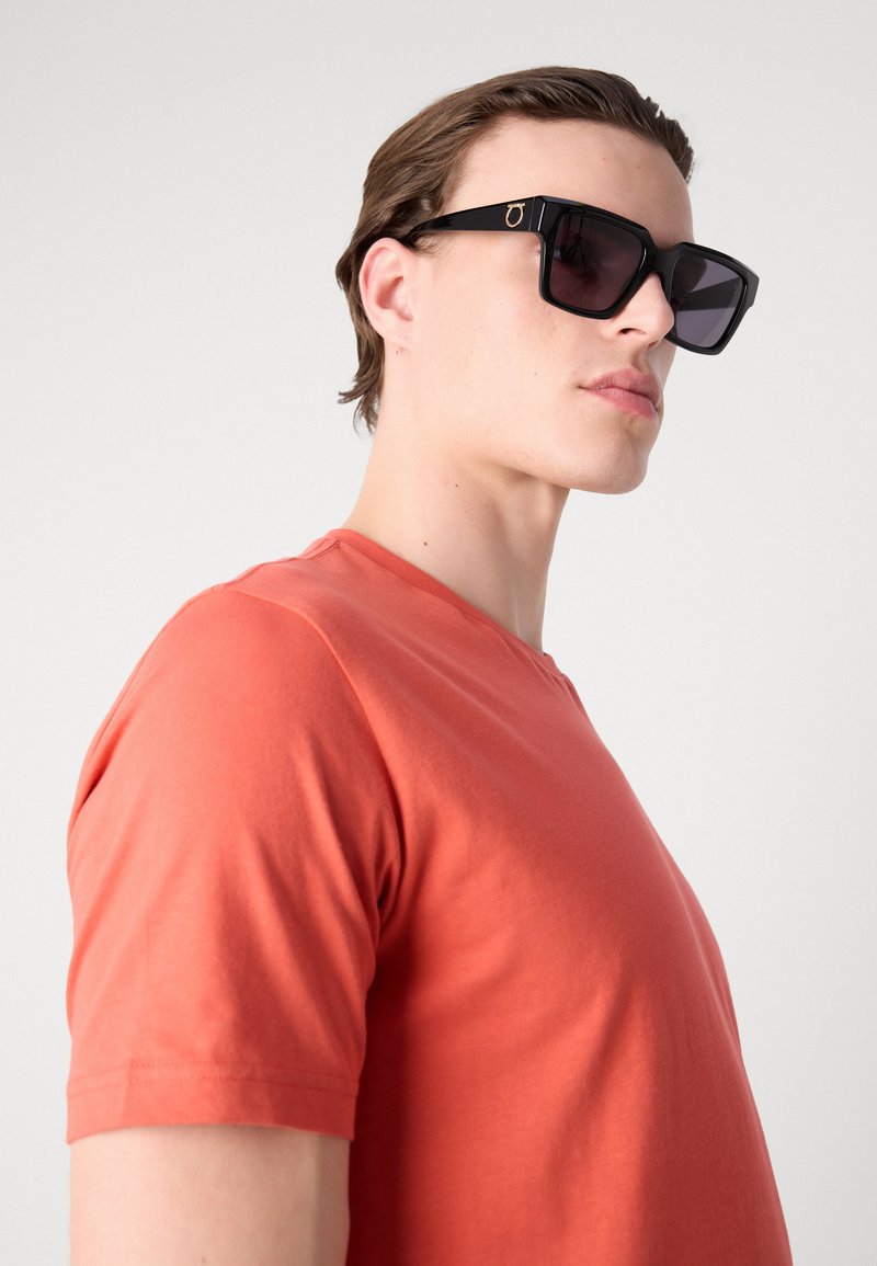 Schwarze rechteckige Sonnenbrillen mit dunklen Gläsern, getragen mit einem kurzärmeligen korallenfarbenen T-Shirt. Glattes Material und elegantes, modernes Design.