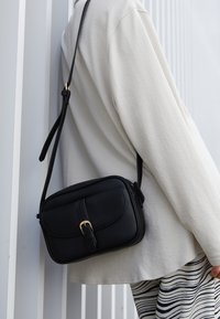 Bolso cruzado de cuero negro con acabado texturizado, detalle de hebilla dorada, forma rectangular y correa ajustable, que se lleva sobre una prenda de color claro.