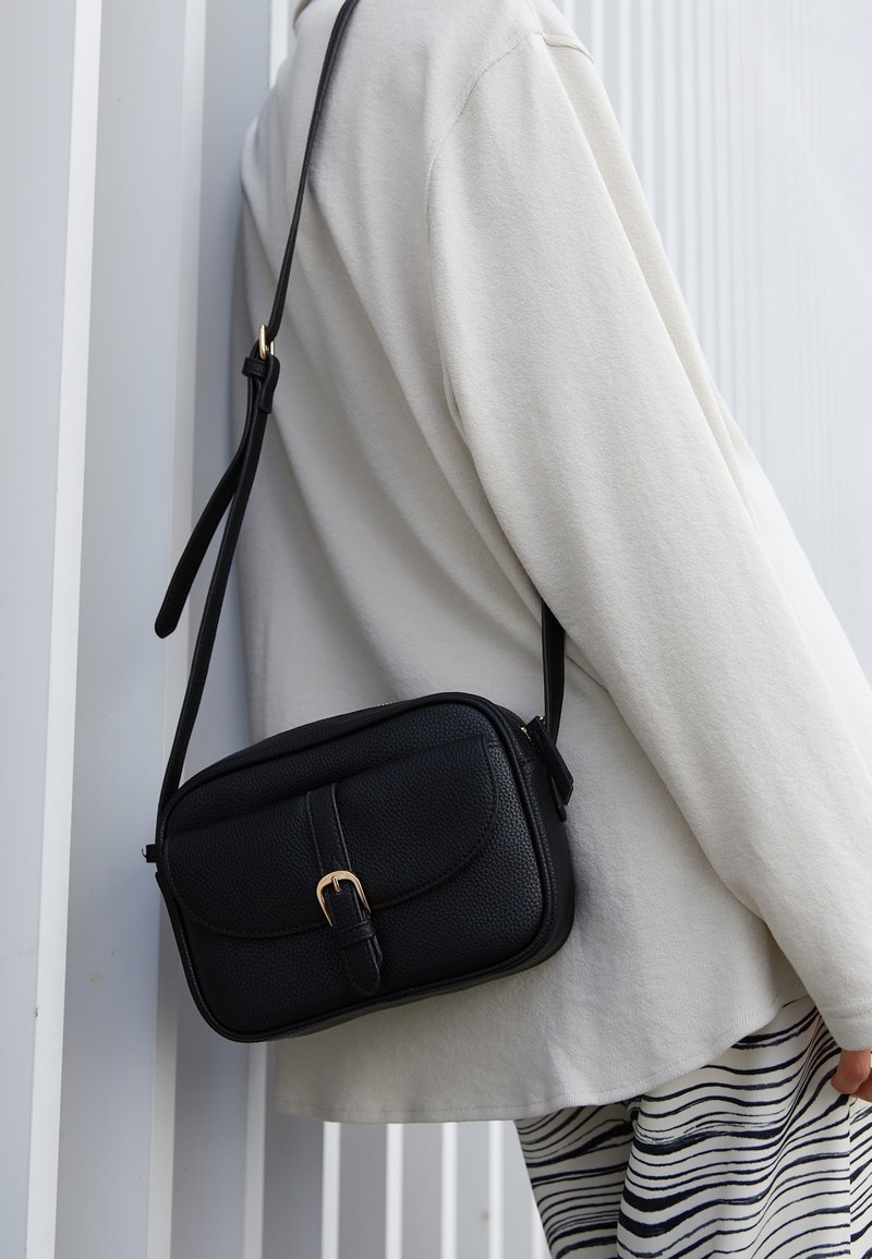 Bolso cruzado de cuero negro con acabado texturizado, detalle de hebilla dorada, forma rectangular y correa ajustable, que se lleva sobre una prenda de color claro.