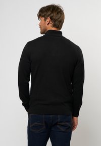 Schwarzer Pullover mit hohem Kragen, langen Ärmeln und gerippten Bündchen. Trägt dazu eine dunkelblaue Jeans mit dekorativen Gesäßtaschen.