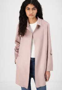 Lys rosa frakk med krage, lange ermer og sidelommer. Glatt tekstur med mansjettaksenter, kombinert med en hvit topp og blå jeans.