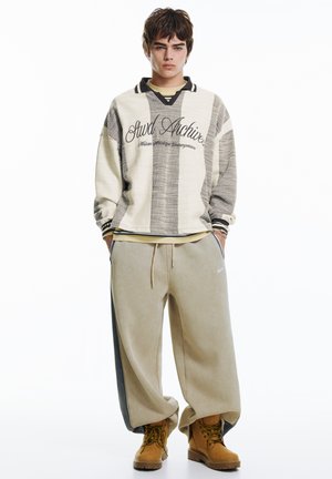 Nadrozmerná béžovo-sivá pruhovaná mikina s vyšitým textom, skombinovaná s voľnými béžovými nohavicami s čiernymi bočnými pruhmi a hnedými čižmami.