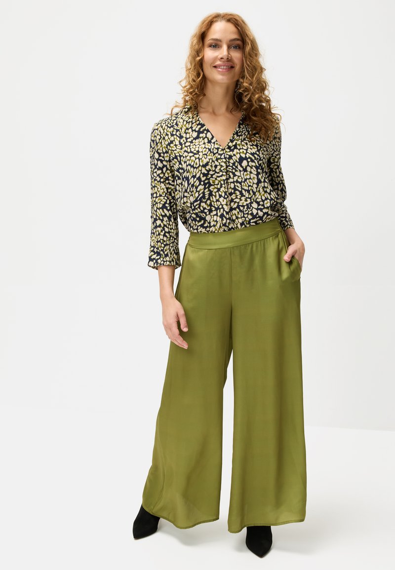 Woman with curly hair wearing a patterned black and yellow blouse tucked into high-waisted olive green wide-leg pants and black shoes, smiling.