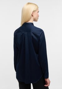 Camicia blu navy a maniche lunghe con texture liscia, colletto su misura e orlo arrotondato. Presenta una plissettatura sul retro e polsini con bottoni.