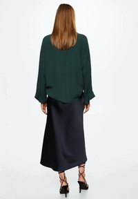 Blouse vert foncé à manches amples associée à une jupe satin noir, longueur cheville, accompagnée de sandales à talons hauts noires avec des lanières entrecroisées.
