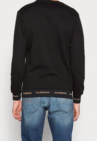 Persona vestido con un sudadera negra con el texto "GLORIOUS" en los puños y el dobladillo, combinada con unos jeans de mezclilla azules, visto desde atrás.