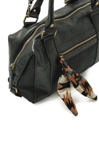 Sac à main en cuir synthétique noir avec des fermetures éclair dorées, deux poignées supérieures et un accent de pompon en tissu à motif. Surface texturée et forme structurée.