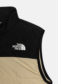 Svart och beige isolerad väst med hög krage, försedd med en framträdande vit logotyp på bröstet och slät textur.