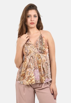 Donna con lunghi capelli castani che indossa un top smanicato a fantasia e pantaloni color malva chiaro su uno sfondo semplice.