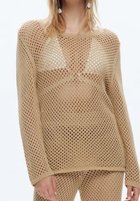 Person wearing beige open-knit long-sleeve top and matching pants with visible white undergarment beneath the top.