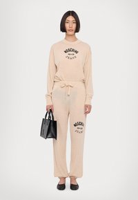 Conjunto de roupa de lazer bege com mangas longas e cintura elástica, apresentando o logo preto "Moschino Jeans". Inclui uma pequena mala tote preta e sapatos pretos planos.