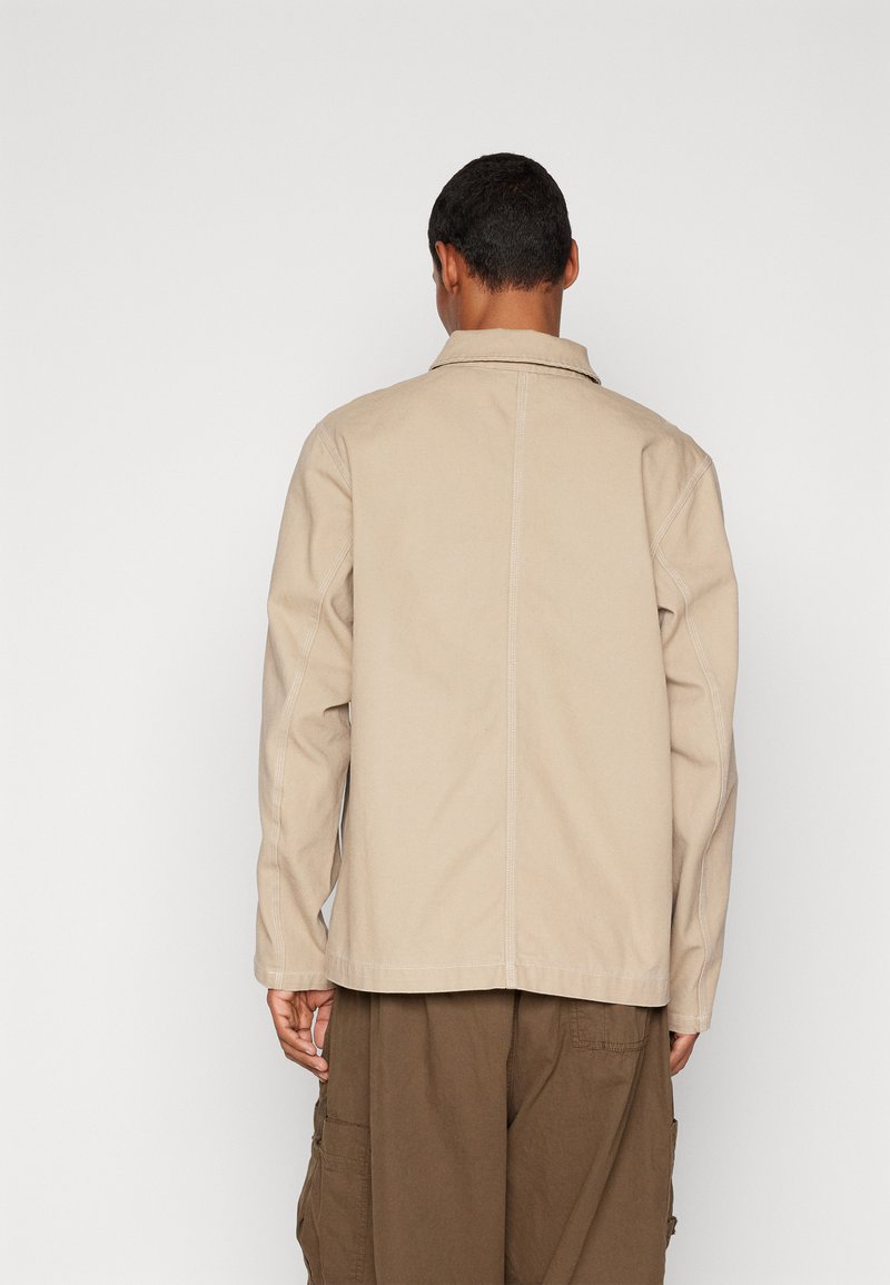 Hombre vistiendo una chaqueta de color beige con cuello y pantalones cargo marrones, de pie con la espalda hacia la cámara contra un fondo blanco liso.