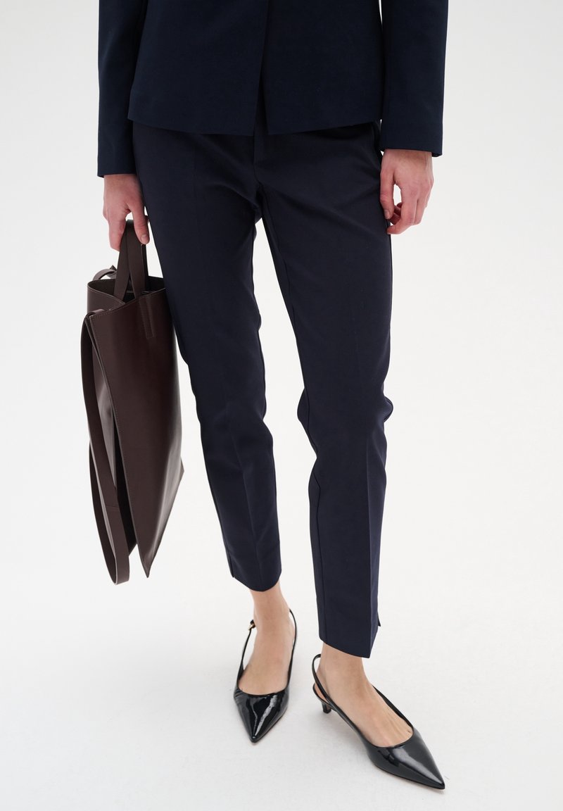 Personne portant un pantalon court bleu marine et des chaussures slingback noires pointues, tenant un grand sac cabas en cuir marron foncé.
