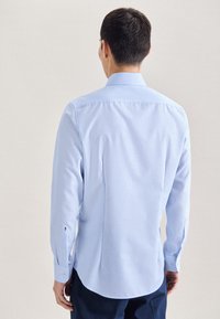 Camicia azzurra chiaro con maniche lunghe, polsini con bottoni, colletto classico e orlo curvo. Tessuto liscio con minima texture.