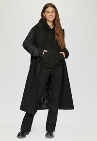 Zwarte lange jas over een zwarte hoodie en broek, met een gestructureerde stof, voorzakken en stevige zwarte laarzen.