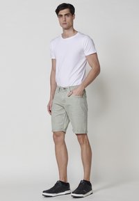 Vyras stovi vilkėdamas baltą trumpą rankovę marškinėlį, šviesiai pilkas trumpikes, suktas iki kelių, ir juodus sportinius batus prieš šviesų vientisą foną.