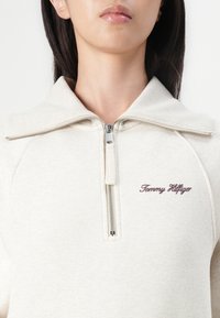 Light beige zip-up sweatshirt featuring a high collar, ribbed details, and "Tommy Hilfiger" logo in burgundy stitching. Soft fabric texture.