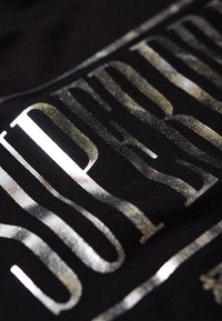 Tissu noir avec du texte métallique argenté présentant une typographie en gras. La texture semble lisse et légèrement réfléchissante.