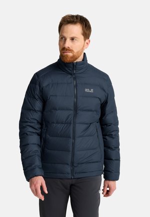 Mann mit rotem Bart, der eine dunkelblaue Steppjacke mit Reißverschlusstaschen trägt und vor einem schlichten Hintergrund steht.