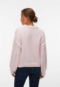 Femme aux cheveux bruns attachés en chignon, portant un pull en maille rose clair avec des manches torsadées et un jean bleu foncé, vue de dos sur un fond blanc.
