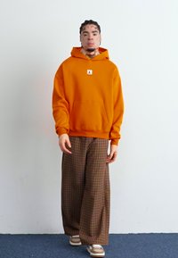 Un sweat à capuche orange avec une poche avant et un logo, associé à un pantalon large à carreaux marron. Les chaussures sont beiges avec des accents blancs sur un sol bleu.