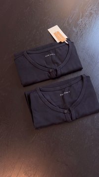 Två vikta svarta T-shirts i mjukt tyg, med rund halsringning, knappdetalj och en etikett synlig på en av tröjorna.