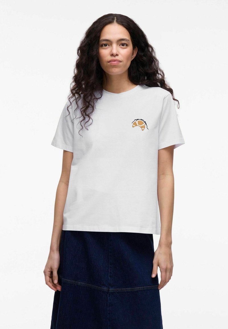 Witte katoenen T-shirt met een croissantafbeelding in bruin en zwarte omlijning op de linkerborst, gecombineerd met een donkerdenim rok.