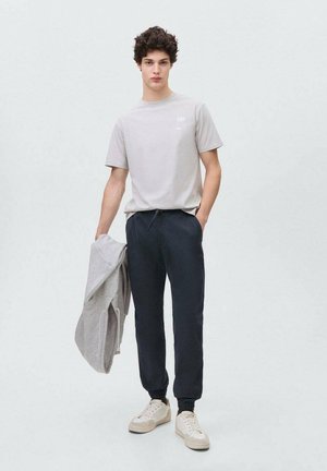 Grå bomulls t-shirt med en liten logotyp, parat med mörka sweatpants och beige sneakers. En ljusgrå sweatshirt hänger över ena armen.