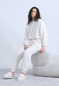 Γυναίκα με σγουρά μαλλιά κάθεται σε καμπυλωτή υφασμάτινη μπεζ καρέκλα, φορώντας λευκό πουλόβερ με ανάγλυφη πλέξη, παντελόνια που ταιριάζουν και παντόφλες σε γκρι χαλί.