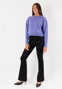 Maglione corto color lavanda con polsini e orlo a costine; pantaloni neri svasati; décolleté nere a punta; logo semplice ricamato sul maglione.