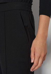 Zwarte broek met een elastische tailleband, geplooid ontwerp en zijzakken. Het materiaal lijkt glad met een lichte textuur.