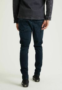 Jeans en denim bleu foncé avec une coupe slim, dotés de deux poches arrière et d'une texture lisse. Associés à des chaussures noires.