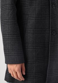 Acercamiento de una mano cerca del bolsillo de un abrigo de lana de cuadros gris oscuro con botones negros, mostrando la textura y el patrón de la tela.