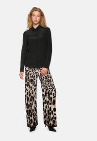 Schwarzes Hemd mit langen Ärmeln, kombiniert mit beigen weiten Hosen, die ein schwarzes Leopardenmuster aufweisen. Schwarze spitze Schuhe.
