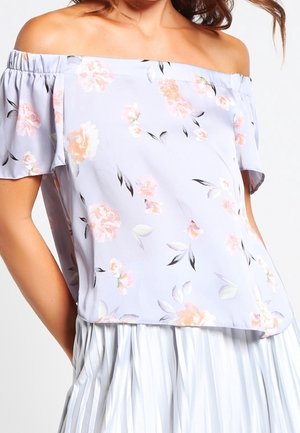 Kvinde iført en lyse lilla off-shoulder bluse med pink blomsterprint og korte ærmer, kombineret med et hvidt plisseret skørt.