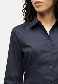 Γυναίκα με κατσαρά μαλλιά, φορώντας στενή σκούρα μπλε πουκαμίσα με κουμπιά, εμφανής από τον λαιμό μέχρι το μέσο του κορμού, με απλό φόντο.