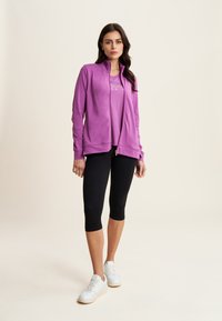 Giacca zip-up viola in morbido tessuto, abbinata a una t-shirt coordinata e leggings capri neri. Scarpe da ginnastica bianche completano l'outfit.
