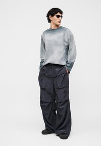 Άνδρας φοράει μαύρα γυαλιά ηλίου, γκρι oversized μακρυμάνικο πουκάμισο και φαρδιά μαύρη cargo παντελόνια με τα χέρια στις τσέπες, στέκεται μπροστά σε μια μονόχρωμη φόντο.