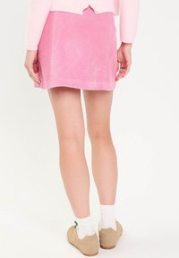 Kobieta w różowej spódniczce z korduroyu, beżowych trampkach, białych skarpetkach i jasnoróżowym swetrze, stoi na tle białej ściany.