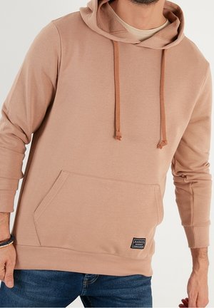 Homme portant un sweat à capuche marron clair avec une poche avant et des cordons, associé à un jean bleu foncé et un bracelet au poignet gauche.