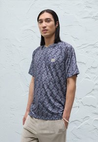 Junger Mann mit langen Haaren, der ein gemustertes graues Sportshirt und hellbeige Hosen trägt, steht vor einer strukturierten cremeweißen Wand.