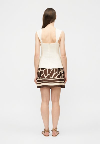 Γυναίκα με μακριά καστανά μαλλιά που φοράει λευκό αμάνικο τοπ και καφέ μικρή φούστα με σχέδια, στεκόμενη πλάτη, φορώντας σανδάλια, σε λευκό φόντο.