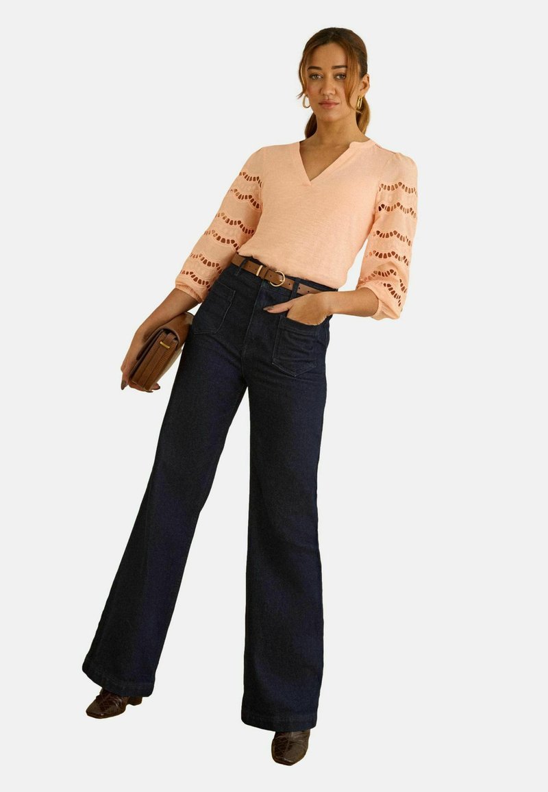 Žena u breskvastoj bluzi s čipkastim rukavima, tamnim širokim trapericama visokog struka, smeđim remenom, smeđom torbicom i smeđim cipelama, stoji i pozira.