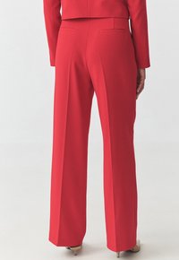 Pantaloni larghi rossi con una texture liscia, dotati di due tasche posteriori e una vestibilità sartoriale, indossati con tacchi in tonalità neutre.