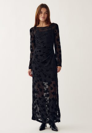 Mujer vestida con un vestido largo negro transparente con patrones florales y mangas largas, combinado con botas negras hasta el tobillo, de pie contra un fondo liso.