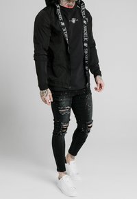 Zwarte hooded jacket met logo-details, zwart t-shirt, gescheurde zwarte jeans met kleurrijke vlekken en witte sneakers.