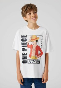 Boy smiling and looking right, wearing a white t-shirt with a red-jacketed anime character holding a straw hat and blue jeans.
