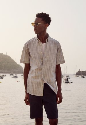 Muškarac obučen u prugastu košulju kratkih rukava, bijelu potkošulju, crne kratke hlače i sunčane naočale stoji kraj vode s čamcima i brežuljcima u pozadini.
