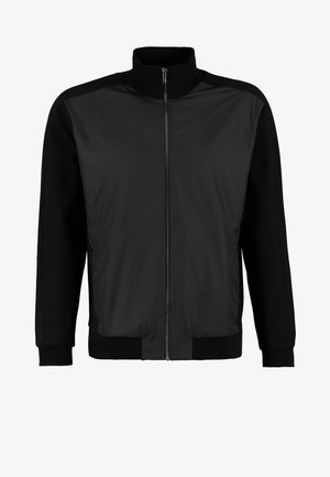 Mann trägt schwarze Jacke, weißes T-Shirt mit großem, schwarz umrandetem Handgrafik, schwarze Hose und schwarze Lederschuhe vor einfachem Hintergrund.